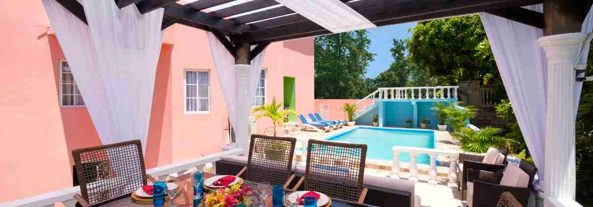 Luxury Villa in Jamaica offers Outdoor dining area with a glass table set for a meal, overlooking a private pool at a peach-colored villa in Jamaica, complete with white sheer curtains and lush greenery in the background